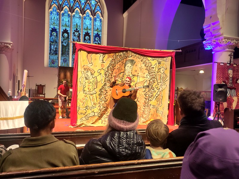 A clown on a Christian altar, in front of a clown backdrop. The clown is holding a guitar.